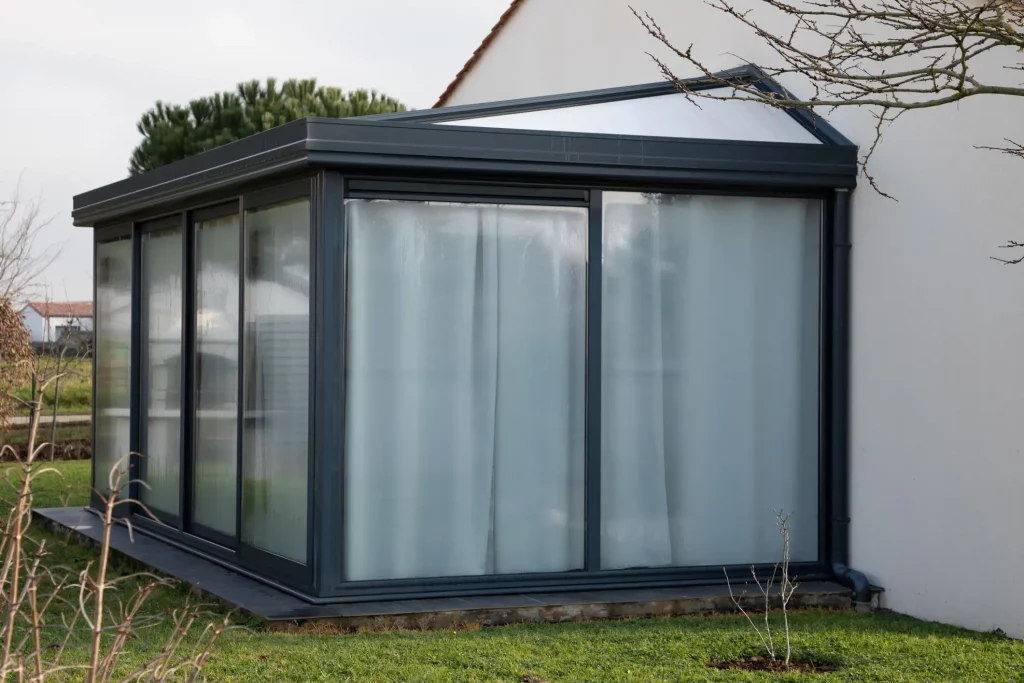 Moderner Wintergartenanbau mit großen Glaswänden, die durch Vorhänge verdeckt sind, an einem Einfamilienhaus, umgeben von einem gepflegten Garten bei bewölktem Himmel.