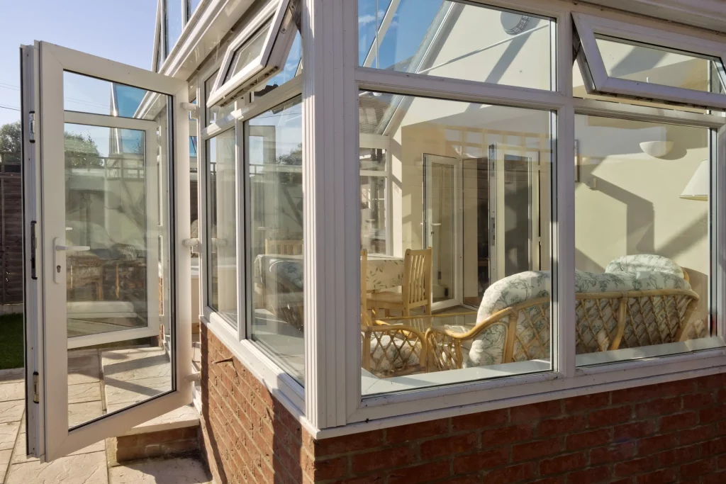 Moderner Wintergarten mit großen Glasfenstern und einer geöffneten Tür, die einen hellen und luftigen Raum mit Korbmöbeln und einem Esstisch im Inneren zeigt. Der Wintergarten ist an ein Haus angeschlossen und bietet einen gemütlichen, lichtdurchfluteten Rückzugsort.