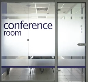 Eine Glastür mit satiniertem Glas eines Konferenzraums mit der Aufschrift ‚conference room‘ in großen Buchstaben. Im Hintergrund sind unscharf Bürostühle und ein Tisch zu erkennen.
