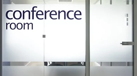 Eine Glastür mit satiniertem Glas eines Konferenzraums mit der Aufschrift ‚conference room‘ in großen Buchstaben. Im Hintergrund sind unscharf Bürostühle und ein Tisch zu erkennen.