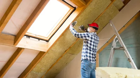 Dachdecker mit roter Kappe und Atemschutzmaske bringt Untersparrendämmung im Dachgeschoss unter einem Dachfenster an. Eine Leiter steht daneben, Holzsparren und Dämmmaterial sind sichtbar.