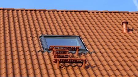 Dachausstiegsfenster auf einem geneigten Dach mit roten Ziegeln und einer kleinen, rot lackierten Trittfläche, die zur Wartung des Daches dient. Im Hintergrund ist ein blauer Himmel sichtbar, und das Fenster ist von Ziegeln sauber eingefasst.