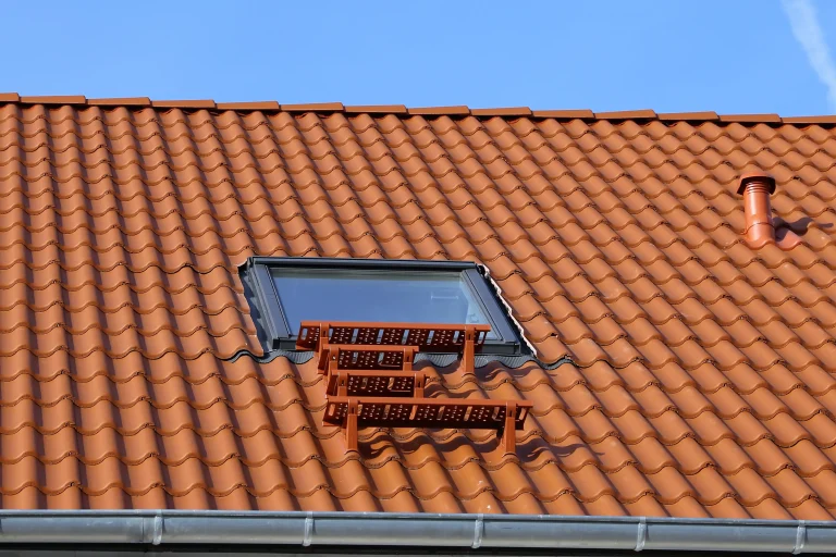 Dachausstiegsfenster auf einem geneigten Dach mit roten Ziegeln und einer kleinen, rot lackierten Trittfläche, die zur Wartung des Daches dient. Im Hintergrund ist ein blauer Himmel sichtbar, und das Fenster ist von Ziegeln sauber eingefasst.