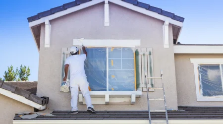 Ein Maler in weißer Arbeitskleidung trägt mit einer Farbrolle Fassadenschutz auf ein Haus auf. Die Fenster sind mit Folie abgedeckt, und der Maler steht auf dem Dach, um die obere Fensterumrandung zu streichen. Eine Leiter lehnt an der Fassade.