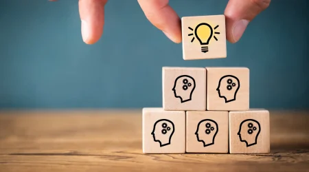 Holzklötze mit Symbolen von Köpfen und Zahnrädern, die für Ideen und Wissen stehen, werden zu einer Pyramide gestapelt. Ein zusätzlicher Klotz mit einer Glühbirne, Symbol für Innovation und neue Ideen, wird auf die Spitze gesetzt.