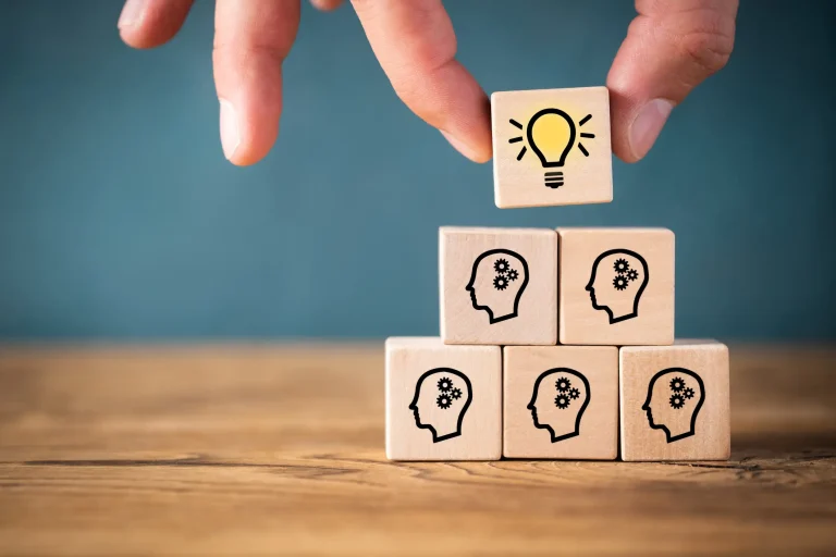 Holzklötze mit Symbolen von Köpfen und Zahnrädern, die für Ideen und Wissen stehen, werden zu einer Pyramide gestapelt. Ein zusätzlicher Klotz mit einer Glühbirne, Symbol für Innovation und neue Ideen, wird auf die Spitze gesetzt.