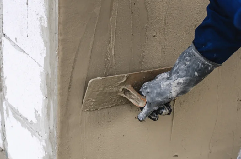 Handwerker trägt Kalkzementputz mit einer Kelle auf eine Außenwand auf. Die Außenwand besteht aus Ziegelsteinen und wird verputzt, um die Oberfläche zu glätten und zu schützen.