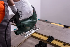 Handwerker benutzt eine Stichsäge zum Schneiden von Laminatboden auf einer Werkbank. Nahaufnahme der Hand mit Schutzhandschuh und der grünen Stichsäge während der Laminatbodenverlegung.