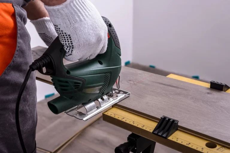 Handwerker benutzt eine Stichsäge zum Schneiden von Laminatboden auf einer Werkbank. Nahaufnahme der Hand mit Schutzhandschuh und der grünen Stichsäge während der Laminatbodenverlegung.
