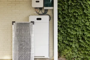 Photovoltaik-Speichersystem an einer Hauswand neben einem neu verpackten Solarmodul, mit grüner, bewachsener Wand im Hintergrund.