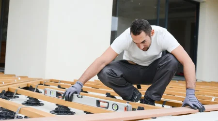 Handwerker beim Bau einer Terrasse, der mit einer Wasserwaage die korrekte Ausrichtung der Unterkonstruktion prüft. Die Terrassendielen sind auf höhenverstellbaren Stellfüßen montiert, um das berechnete Gefälle zu erreichen. Durch die Neigung soll gewährleistet werden, dass Regenwasser vom Haus weg abfließt.