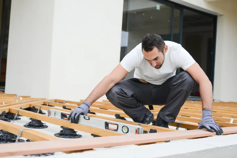 Handwerker beim Bau einer Terrasse, der mit einer Wasserwaage die korrekte Ausrichtung der Unterkonstruktion prüft. Die Terrassendielen sind auf höhenverstellbaren Stellfüßen montiert, um das berechnete Gefälle zu erreichen. Durch die Neigung soll gewährleistet werden, dass Regenwasser vom Haus weg abfließt.