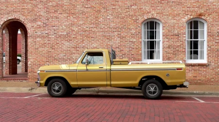 Goldfarbener Pickup-Truck der Ford F-Serie aus den 1970er Jahren, geparkt auf rotem Kopfsteinpflaster vor einem historischen Ziegelbau mit weißen Fenstern.