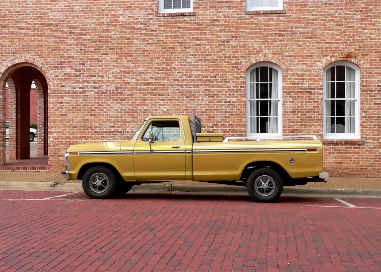 Goldfarbener Pickup-Truck der Ford F-Serie aus den 1970er Jahren, geparkt auf rotem Kopfsteinpflaster vor einem historischen Ziegelbau mit weißen Fenstern.