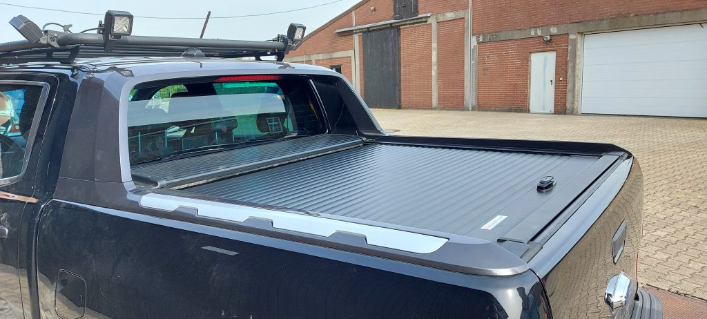 Schwarzer Pickup mit geschlossener Aluminium-Rollladenabdeckung und Dachgepäckträger mit LED-Arbeitsscheinwerfern, geparkt auf einem gepflasterten Industriehof vor einem Ziegelgebäude.