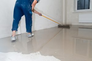 Handwerker in blauer Arbeitskleidung verteilt Anhydritestrich mit einem Rakel auf einem Boden in einem leeren Raum. Glatte Oberfläche des Estrichs spiegelt Licht wider, während der Handwerker für eine gleichmäßige Verteilung sorgt.
