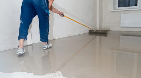 Handwerker in blauer Arbeitskleidung verteilt Anhydritestrich mit einem Rakel auf einem Boden in einem leeren Raum. Glatte Oberfläche des Estrichs spiegelt Licht wider, während der Handwerker für eine gleichmäßige Verteilung sorgt.