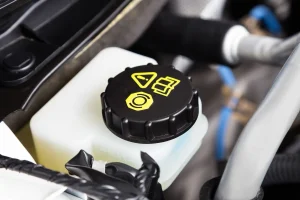 Verschlussdeckel des Bremsflüssigkeitsbehälters in einem Fahrzeugmotorraum, mit gelben Symbolen zur Warnung und Identifikation. Der schwarze Deckel sitzt auf einem weißen Kunststoffbehälter, der den Füllstand der Bremsflüssigkeit anzeigt.
