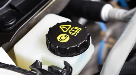 Verschlussdeckel des Bremsflüssigkeitsbehälters in einem Fahrzeugmotorraum, mit gelben Symbolen zur Warnung und Identifikation. Der schwarze Deckel sitzt auf einem weißen Kunststoffbehälter, der den Füllstand der Bremsflüssigkeit anzeigt.