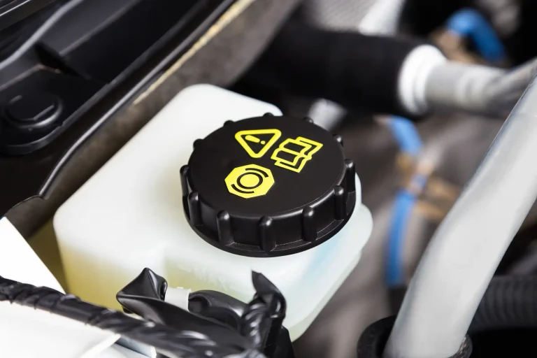 Verschlussdeckel des Bremsflüssigkeitsbehälters in einem Fahrzeugmotorraum, mit gelben Symbolen zur Warnung und Identifikation. Der schwarze Deckel sitzt auf einem weißen Kunststoffbehälter, der den Füllstand der Bremsflüssigkeit anzeigt.