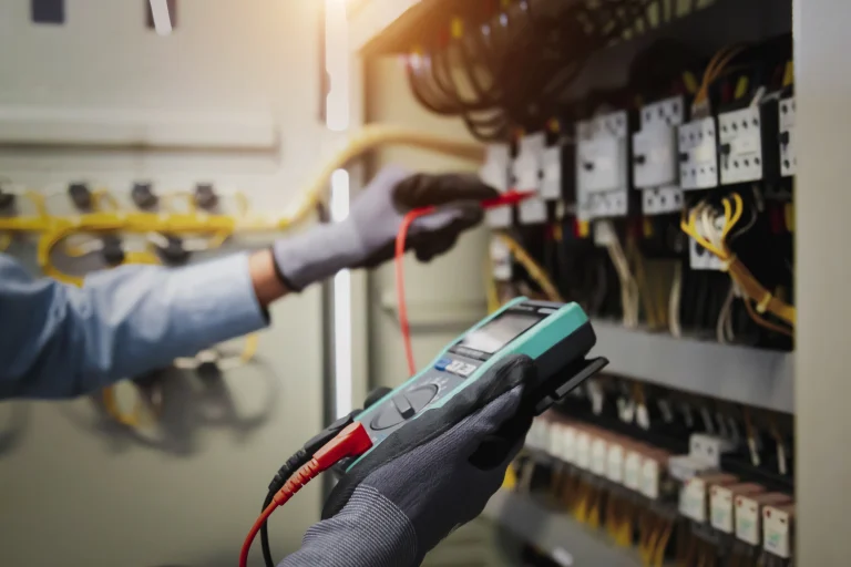 Elektriker misst mit einem Multimeter die Spannung in einem Schaltschrank. Zahlreiche Kabel und Schalterelemente sind sichtbar, die Hände sind mit Schutzhandschuhen gesichert.