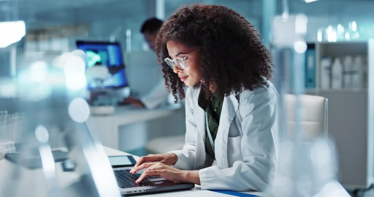 Junge Wissenschaftlerin im Labor, konzentriert am Laptop arbeitend, um Forschung und Innovation voranzutreiben. Im Hintergrund moderne Laborgeräte und Computerbildschirme.