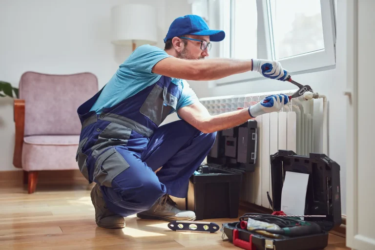 Ein Heizungsbauer in blauer Arbeitskleidung entlüftet eine pfeifende Heizung in einem Wohnzimmer. Er verwendet eine Zange, während verschiedene Werkzeuge in einem offenen Werkzeugkoffer neben ihm auf dem Boden liegen.
