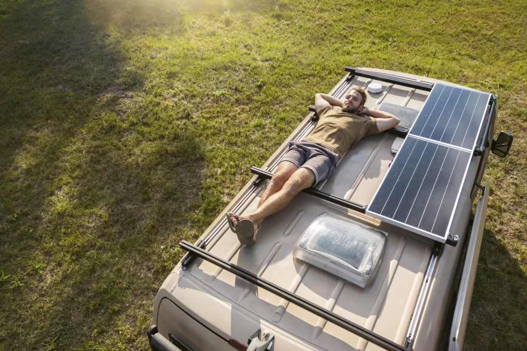 Mann entspannt sich auf dem Dach eines Campervans, der mit einer netzunabhängigen Photovoltaikanlage ausgestattet ist. Die Solarzellen auf dem Fahrzeugdach ermöglichen autarke Stromversorgung für mobiles Reisen und Camping. 
