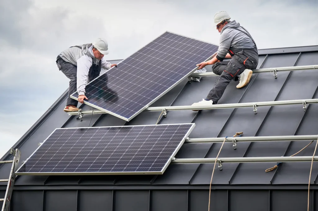 Zwei Handwerker montieren Photovoltaik-Module auf einem geneigten Metalldach. Beide tragen Schutzhelme und arbeiten sorgfältig an der Installation der Photovoltaikanlage.