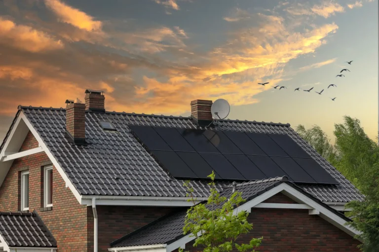 Einfamilienhaus mit modernem Ziegeldach, ausgestattet mit Photovoltaik-Modulen zur Solarstromerzeugung. Das Haus verfügt über zwei Schornsteine und eine Satellitenschüssel, umgeben von grünen Bäumen bei Sonnenuntergang.