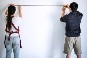 Ein Paar misst gemeinsam eine weiße Wand mit einem Maßband aus, um den Tapetenbedarf zu berechnen. Die Frau trägt eine weiße Bluse und eine rote Schürze, während der Mann ein dunkelblaues Hemd und beige Shorts trägt.