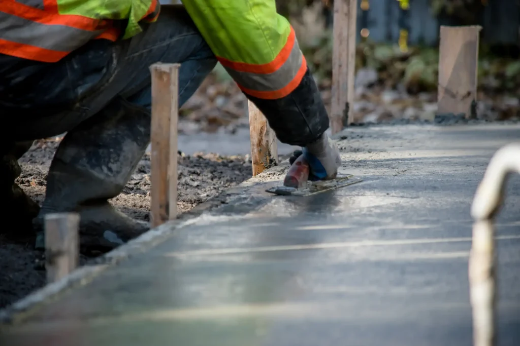 Handwerker in reflektierender Arbeitskleidung glättet mit einer Kelle frisch gegossenen Beton für das Fundament eines Wintergartens.