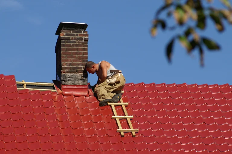 Handwerker auf einem roten Metalldach, der an einem alten Ziegelsteinkamin arbeitet. Der Kamin ist mit einer Metallabdichtung gesichert, und der Arbeiter nutzt eine kleine Holzleiter.
