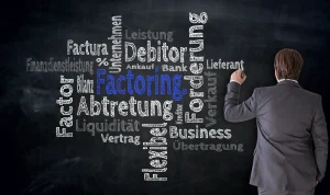 Ein Geschäftsmann in einem grauen Anzug steht vor einer schwarzen Tafel und schreibt mit einem Marker. Auf der Tafel sind verschiedene Begriffe in deutscher Sprache zu sehen, die sich auf Factoring und Finanzdienstleistungen beziehen.