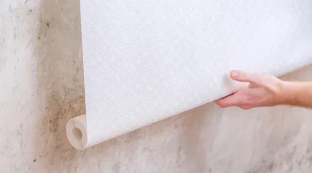 Eine Person hält eine Rolle weiße Fasertapete und bereitet sich darauf vor, diese an einer Wand mit rauer, fleckiger Oberfläche anzubringen.