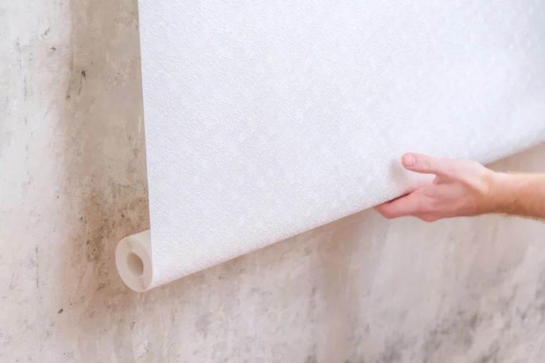 Eine Person hält eine Rolle weiße Fasertapete und bereitet sich darauf vor, diese an einer Wand mit rauer, fleckiger Oberfläche anzubringen.