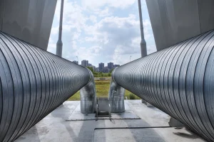 Große, silberne Fernleitungsrohre mit einer gerippten Metalloberfläche verlaufen parallel und biegen sich in der Mitte nach unten. Sie sind auf einer Betonplattform montiert. Im Hintergrund sind eine Wiese, eine Straße mit Straßenlaternen und moderne Hochhäuser unter einem bewölkten Himmel zu sehen.