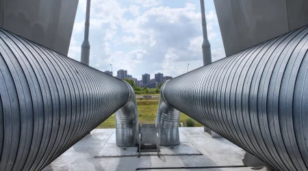 Große, silberne Fernleitungsrohre mit einer gerippten Metalloberfläche verlaufen parallel und biegen sich in der Mitte nach unten. Sie sind auf einer Betonplattform montiert. Im Hintergrund sind eine Wiese, eine Straße mit Straßenlaternen und moderne Hochhäuser unter einem bewölkten Himmel zu sehen.