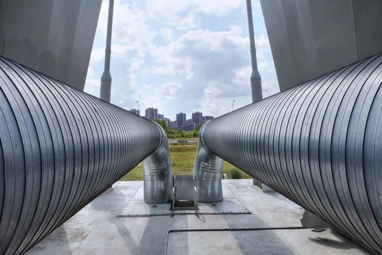 Große, silberne Fernleitungsrohre mit einer gerippten Metalloberfläche verlaufen parallel und biegen sich in der Mitte nach unten. Sie sind auf einer Betonplattform montiert. Im Hintergrund sind eine Wiese, eine Straße mit Straßenlaternen und moderne Hochhäuser unter einem bewölkten Himmel zu sehen.