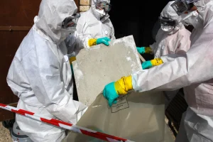 Vier Personen in weißen Schutzanzügen, Schutzbrillen, Atemmasken und grünen Handschuhen tragen eine rechteckige Asbestplatte mit abgenutzter Oberfläche. Sie halten die Platte über einen großen, gekennzeichneten Kunststoffsack. Eine rot-weiße Absperrung ist im Vordergrund sichtbar.