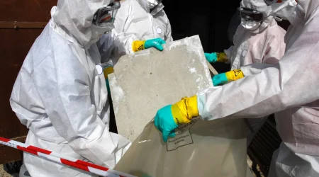 Vier Personen in weißen Schutzanzügen, Schutzbrillen, Atemmasken und grünen Handschuhen tragen eine rechteckige Asbestplatte mit abgenutzter Oberfläche. Sie halten die Platte über einen großen, gekennzeichneten Kunststoffsack. Eine rot-weiße Absperrung ist im Vordergrund sichtbar.