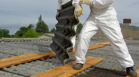 Zwei Handwerker in Schutzanzügen und Handschuhen entfernen eine wellige Asbest-Dachplatte von einem alten Gebäude. Sie stehen auf Holzplanken, um die brüchige Oberfläche des Dachs nicht zu beschädigen. Die Umgebung zeigt eine ländliche Landschaft mit grünen Bäumen und einem leicht bewölkten Himmel.