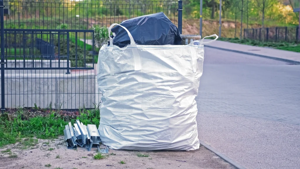 Großer weißer Big Bag mit Trageschlaufen, gefüllt mit Asbest, steht auf einem sandigen Untergrund neben einem Metallzaun. Oben ragt eine schwarze Folie aus dem Sack. Daneben liegt ein Stapel verzinkter Metallprofile auf dem Boden. Im Hintergrund eine gepflasterte Straße und eine Grünanlage.
