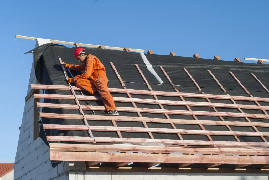Dachdecker in orangem Arbeitsanzug und rotem Schutzhelm arbeitet bei einer Dachsanierung auf einem Hausdach mit Dachlatten und Abdichtungsfolie