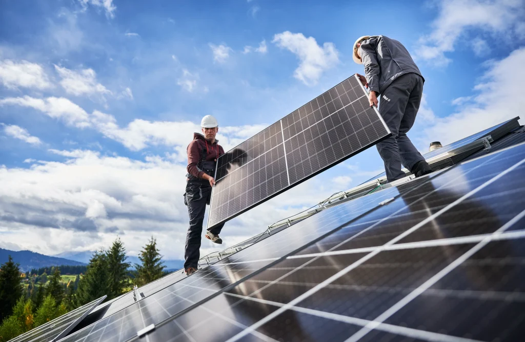 Zwei Handwerker installieren Photovoltaikmodule auf einem schrägen Dach. Beide tragen Schutzhelme und Arbeitskleidung.