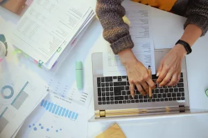 Person tippt auf einem Laptop, umgeben von Finanzunterlagen, Diagrammen und einem Textmarker. Symbolbild digitaler Buchhaltung.