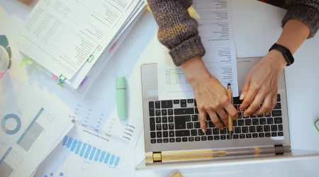 Person tippt auf einem Laptop, umgeben von Finanzunterlagen, Diagrammen und einem Textmarker. Symbolbild digitaler Buchhaltung.