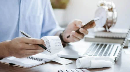 Eine Person in einem blauen Hemd prüft Belege, hält einen Stift und ein Smartphone in den Händen. Im Vordergrund befinden sich ein Taschenrechner, ein Notizbuch und weitere Belege, während im Hintergrund ein Laptop und eine Uhr sichtbar sind.