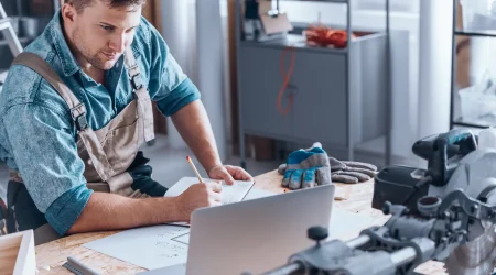 Handwerker in Arbeitskleidung sitzt an einer Werkbank mit Laptop, Bauplänen und Notizbuch. Er macht sich Notizen und informiert sich über Facotring im Handwerk, während eine Kreissäge und Arbeitshandschuhe im Vordergrund liegen.
