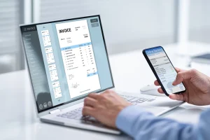 Ein Laptop mit einer geöffneten Rechnungsverwaltungssoftware zeigt eine digitale Rechnung. Eine Hand hält ein Smartphone mit einer Online-Banking-App.